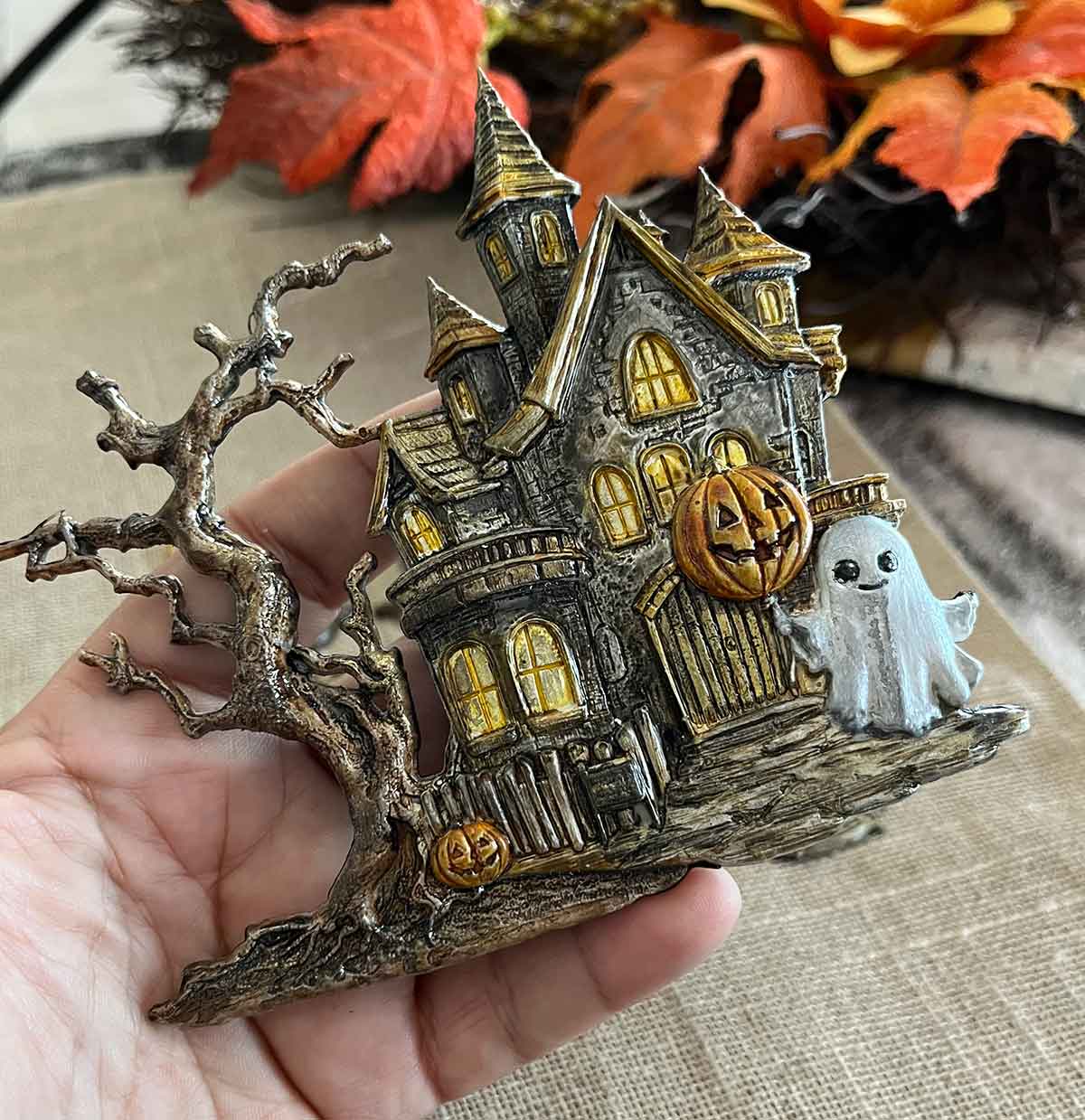 Halloween-themed miniature house with ghost and pumpkin on a hand, with autumn leaves in the background.