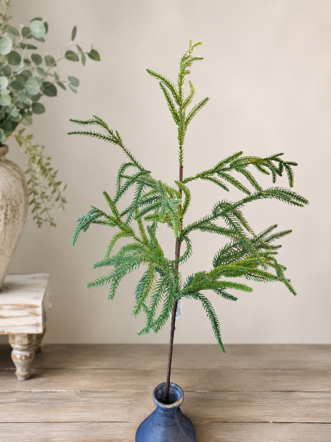 Blue vase with 1 Norfolk pine pick with a white wall in the background