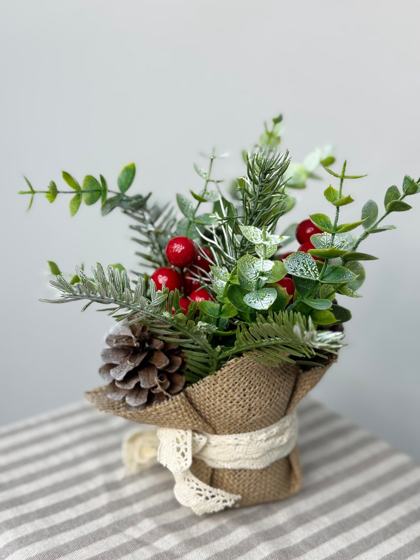 the side view-close up of small eucalyptus arrangement in a burlap base. It has pinecones, cedar, pine branches, and red berries.