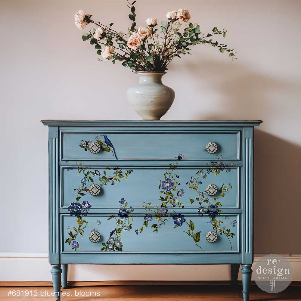 Blue dresser with floral designs and a vase of flowers on top against a plain wall.