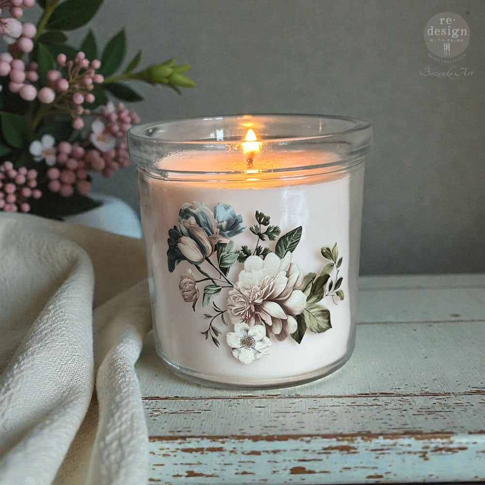 Candle in a glass jar with floral design on a wooden surface, next to pink flowers.