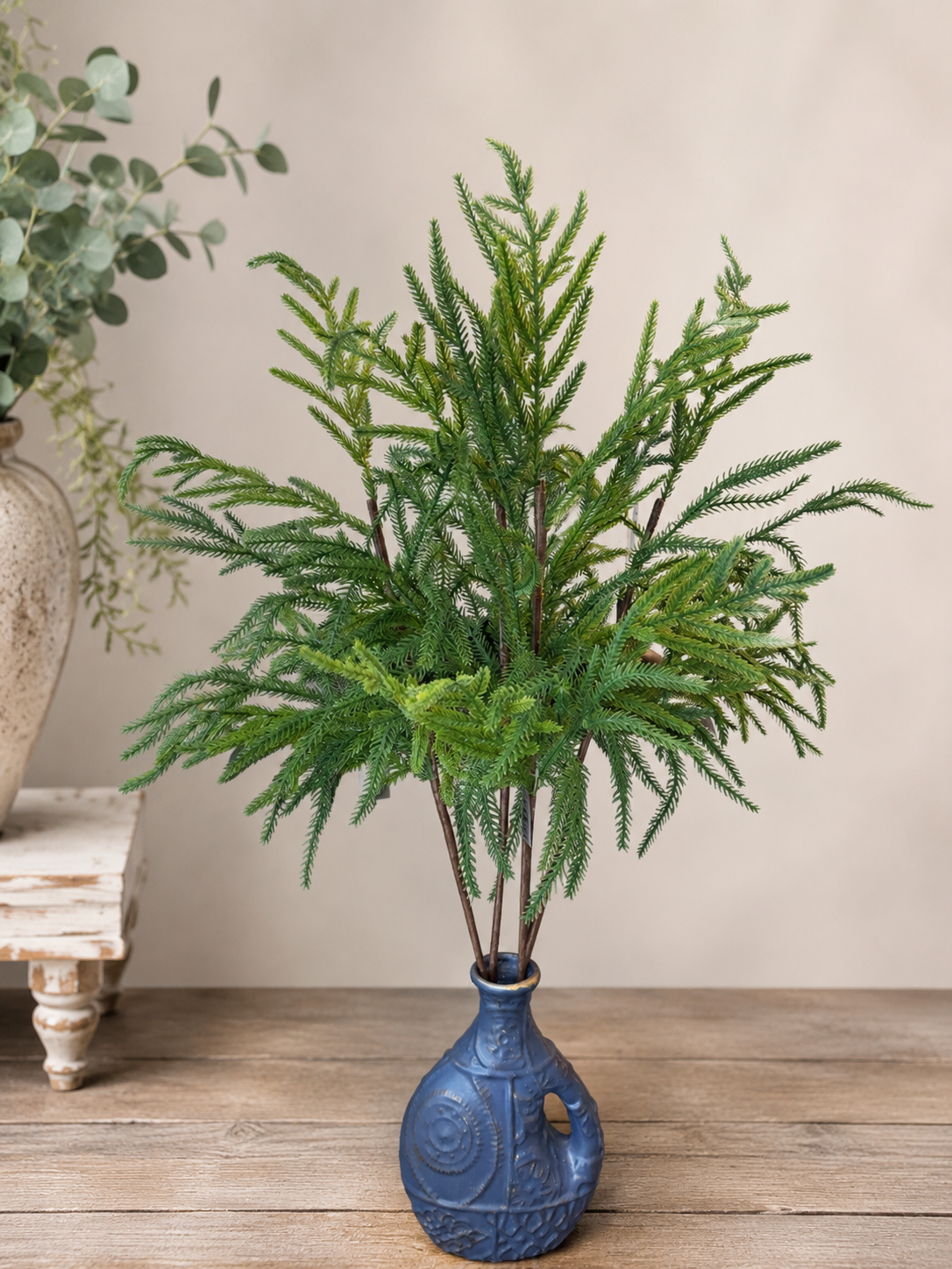 Blue vase with 4 Norfolk pine picks with a white wall in the background