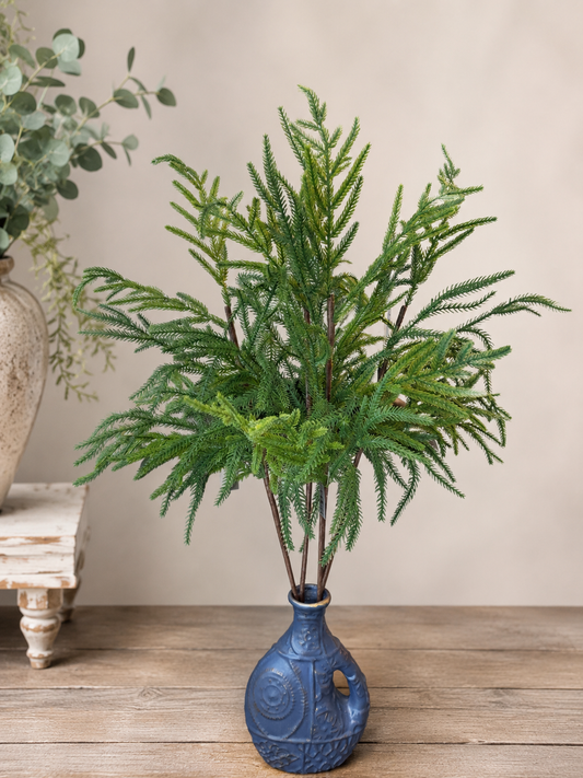 Blue vase with 4 Norfolk pine picks with a white wall in the background