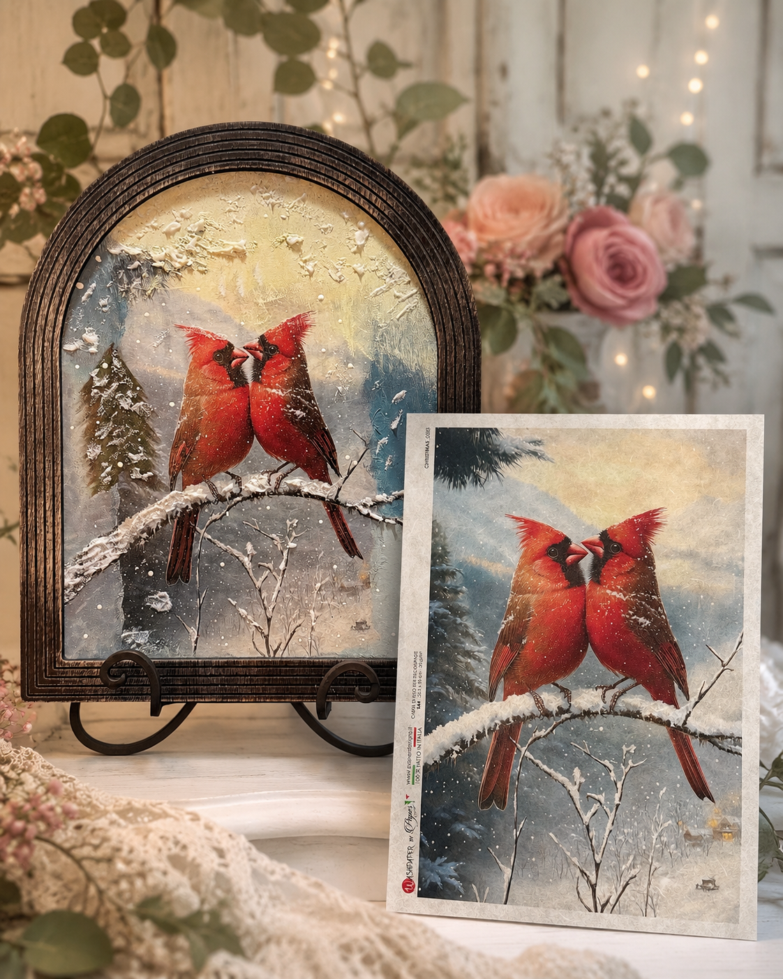 A framed art print and matching greeting card featuring two red cardinals perched on a snowy branch, styled with flowers and greenery in a warm indoor setting.
