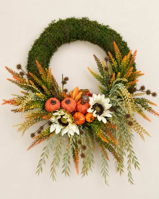 An autumn-themed moss wreath decorated with faux pumpkins, white and orange flowers, and green and orange foliage, set against a plain light beige background.