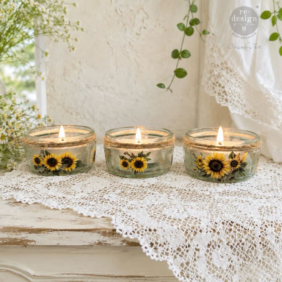 Three glass candles with sunflower designs on a lace tablecloth.