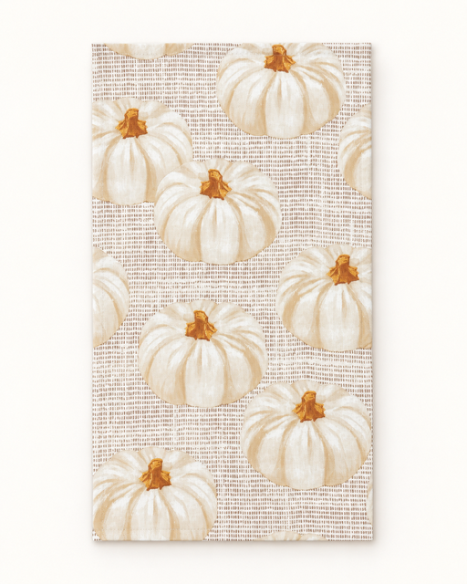 A rectangular fabric item, likely a tea towel or napkin, featuring a repeating pattern of white pumpkins with orange stems on a textured beige woven background.