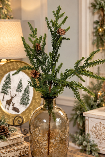 Artificial green pine branches with small pinecones arranged in a clear glass vase with a gold-toned base, set in a warmly lit holiday room setting.