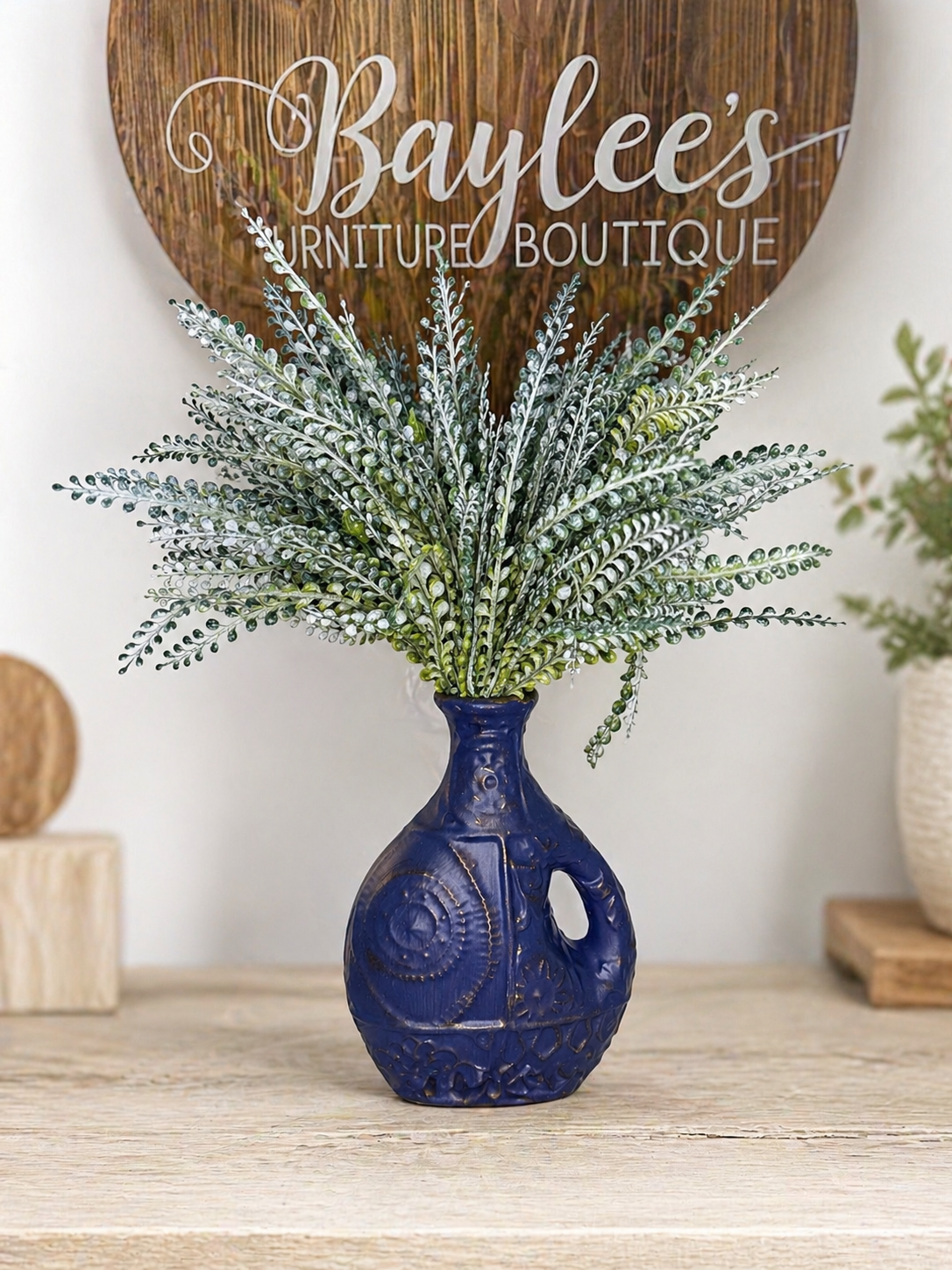 Navy blue vase with greenery on a wooden surface, with 'Baylee's Furniture Boutique' sign in the background.