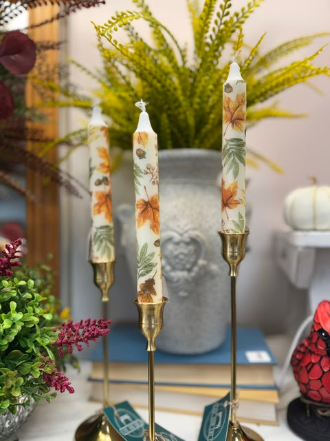 Three taper candles with an autumn leaf pattern in orange, brown, and green, displayed in brass candlesticks on a tabletop with decorative plants and books in the background.