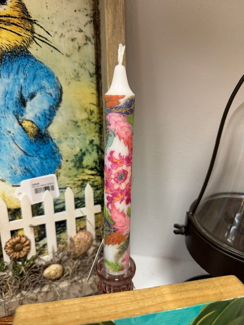 A tall decorative candle with a colorful floral print wrapped around its body, standing upright on a small brown base. The candle has a white wax top and is photographed in a styled indoor setting with a painting, small decorative objects, and a glass dome in the background.