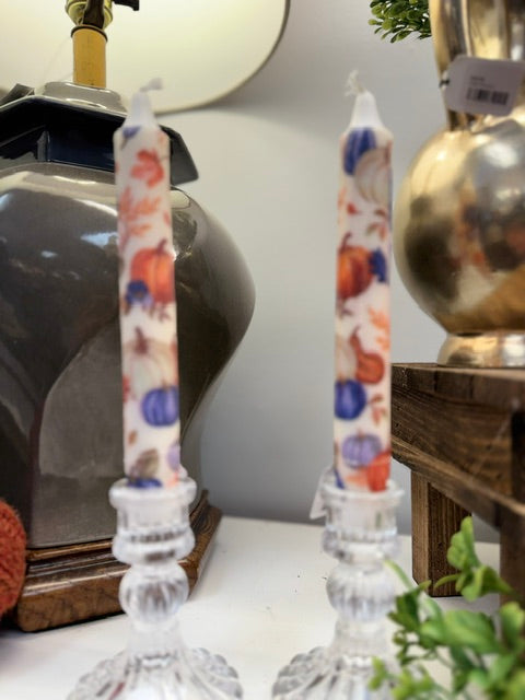 Two white taper candles with a colorful autumn pumpkin and gourd pattern, displayed in clear glass candle holders on a tabletop.