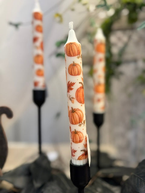A white taper candle with an orange pumpkin and autumn leaf pattern, displayed in a black metal holder. Two matching candles are softly blurred in the background.