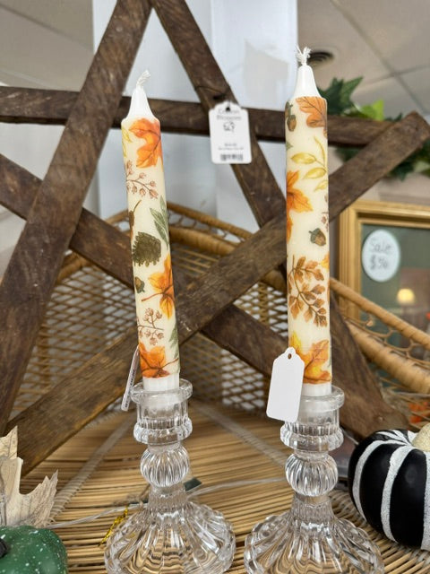 Two taper candles with an autumn leaf pattern, displayed in clear glass candle holders on a woven mat with rustic wooden decor in the background.
