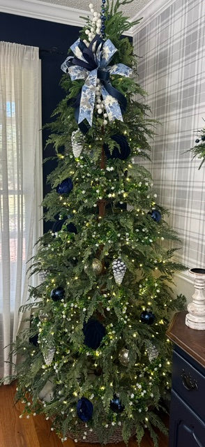 A decorated Christmas tree in a home interior, featuring green branches with white lights, navy blue ornaments, and a large navy and white bow at the top.