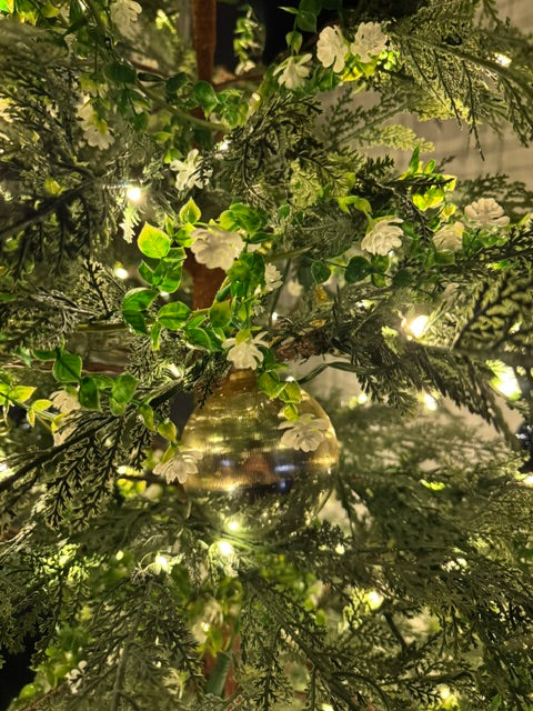 Close-up of a decorated artificial Christmas tree with green faux foliage, small white flowers, warm white string lights, and a large clear round ornament reflecting the room.