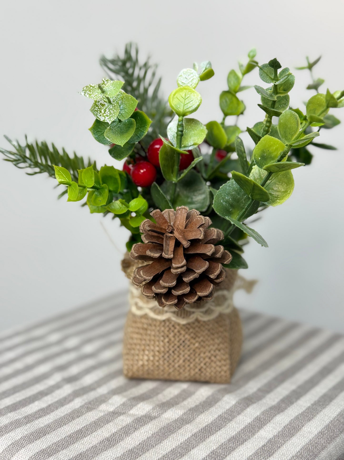 the back-close up of small eucalyptus arrangement in a burlap base. It has pinecones, cedar, pine branches, and red berries.
