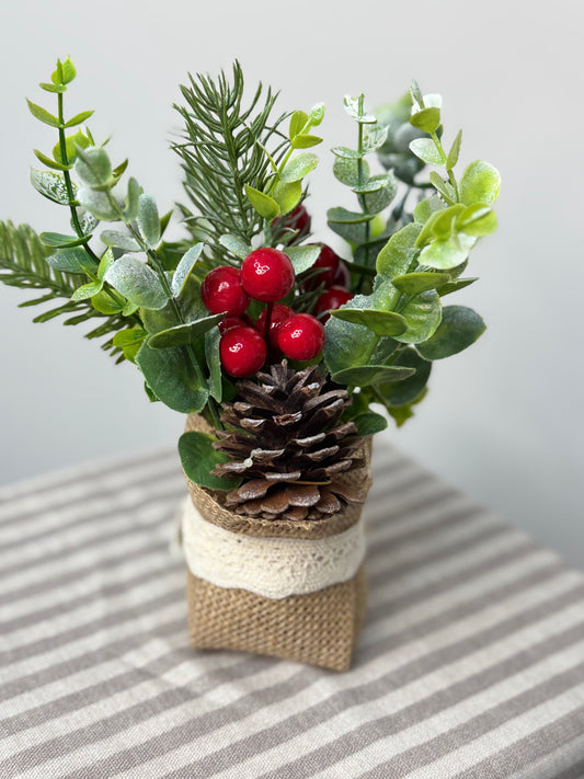 the back view-close up of small eucalyptus arrangement in a burlap base. It has pinecones, cedar, pine branches, and red berries.
