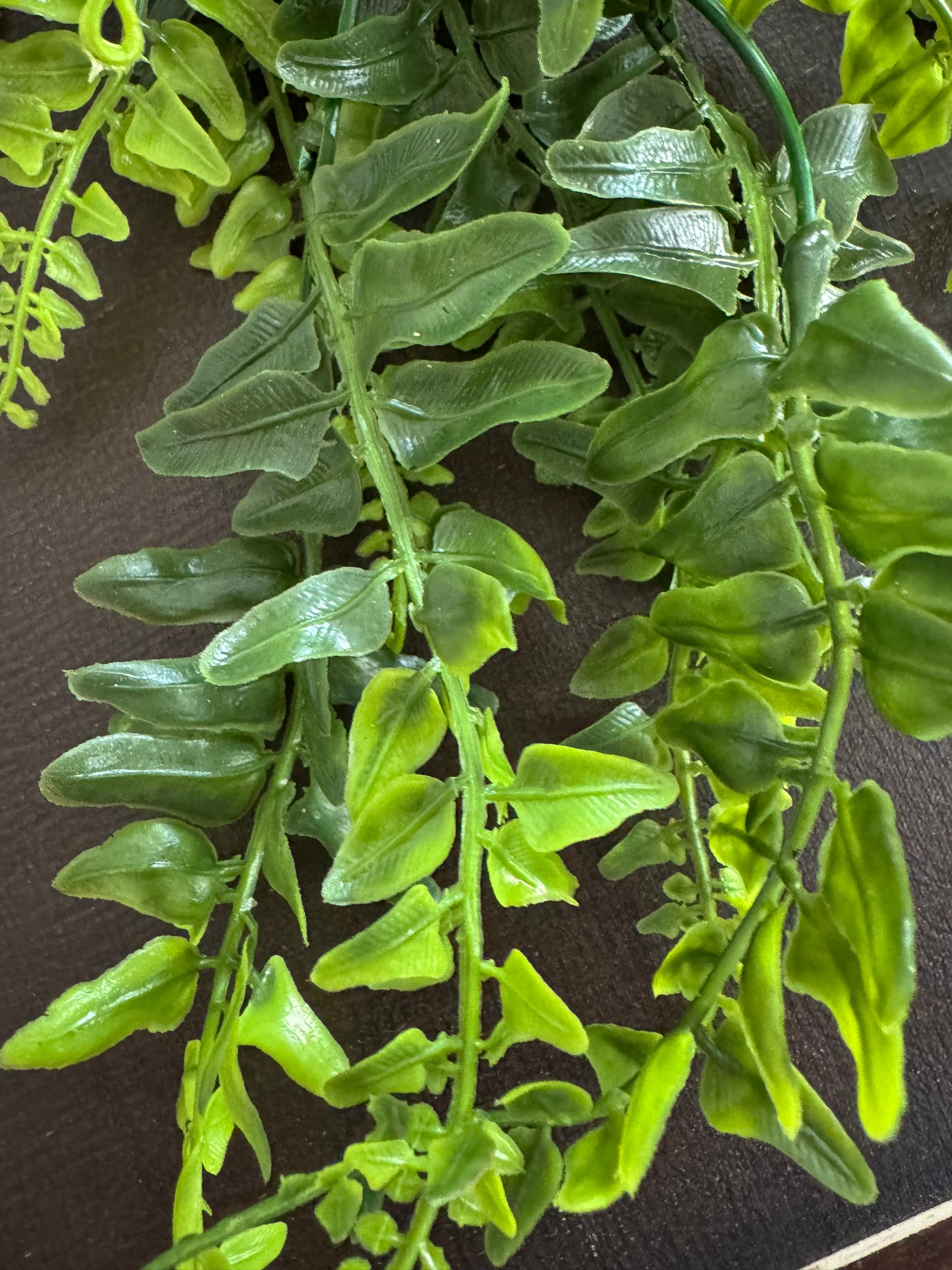 Green artificial fern foliage with realistic textured leaves on a dark brown background.