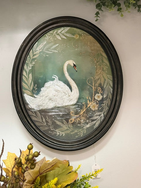 An oval framed wall art piece featuring a white swan painted on a muted green background with botanical leafy details and a dark ornate frame, displayed against a light wall with decorative foliage around it.