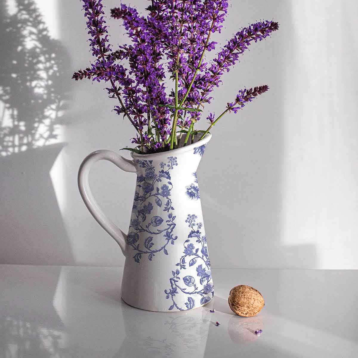 White pitcher with blue floral patterns filled with purple flowers on a white surface.