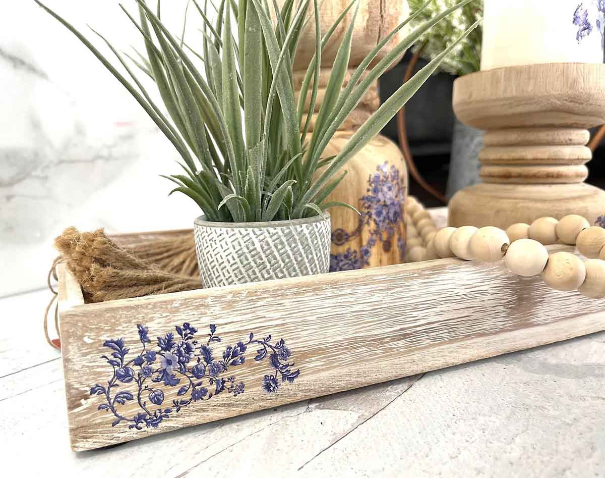 Decorative wooden tray with a plant, beads, and a candle on a light surface.