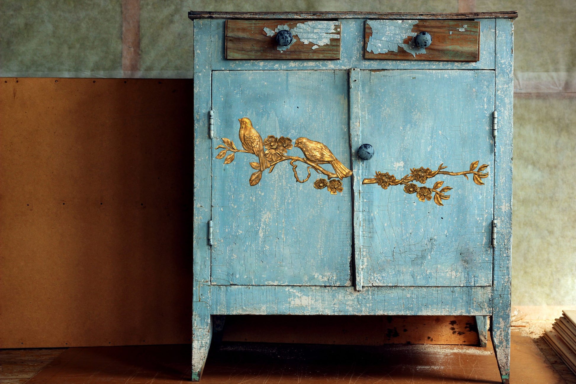 Vintage blue cabinet with gold floral designs against a textured wall.