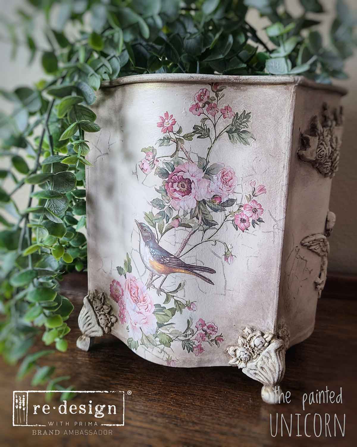 Decorative planter with floral and bird design on a wooden surface with green plants.