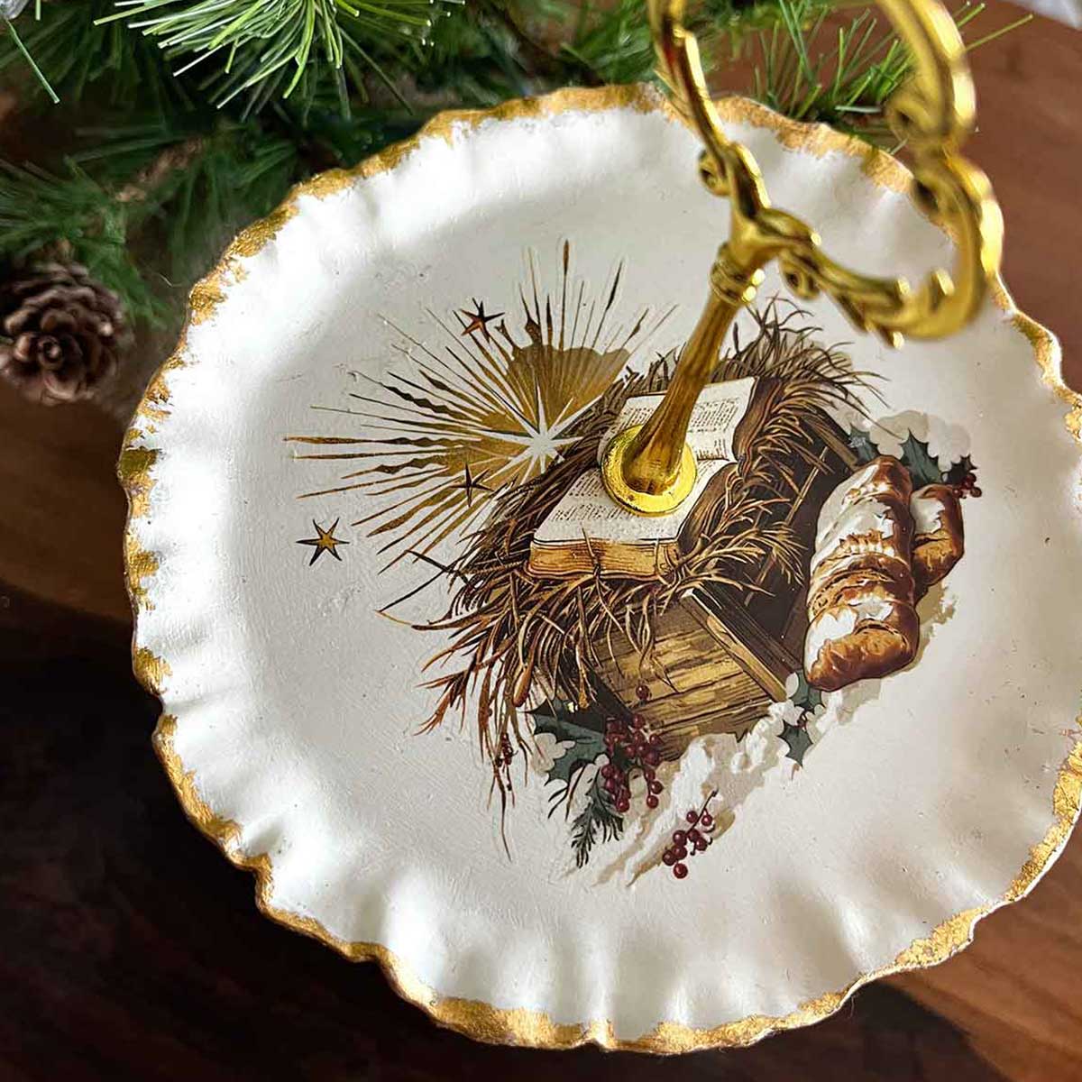 Decorative plate with Christmas scene and gold rim, hanging on a tree.