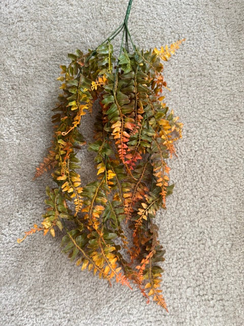 Artificial autumn fern garland with green, yellow, and orange faux leaves hanging against a textured gray carpet background.