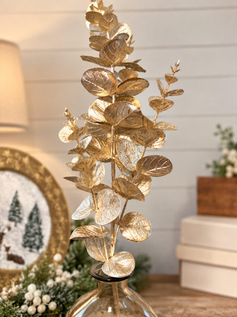 A decorative artificial eucalyptus-style branch with metallic gold leaves, shown in a softly blurred indoor setting with a light wall and tabletop visible.