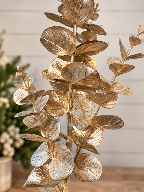 A decorative artificial eucalyptus-style branch with metallic gold leaves, shown in a softly blurred indoor setting with a light wall and tabletop visible.