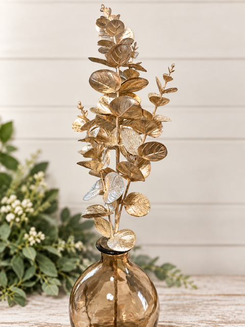A decorative artificial eucalyptus branch with metallic gold leaves arranged in a rounded amber glass vase, set against a light wood paneled background with blurred green foliage on the left.