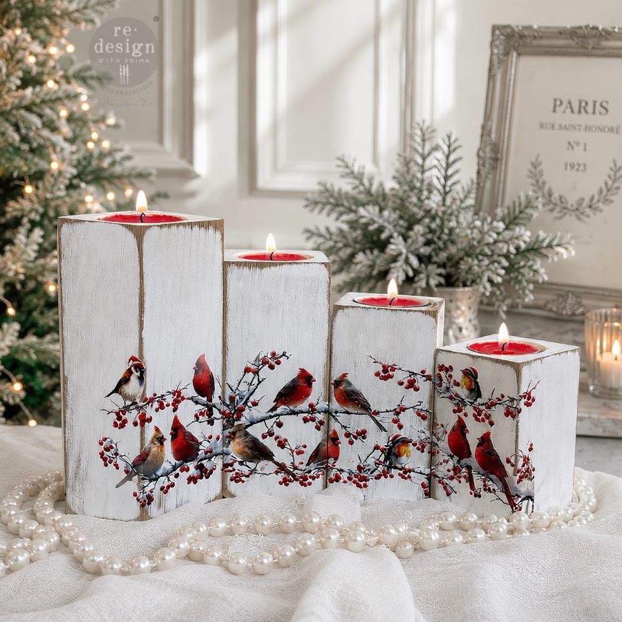 Decorative candles with bird and berry design on a white surface with Christmas decorations in the background.